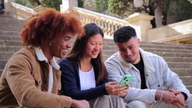 Çok ırklı bir grup genç arkadaş cep telefonu uygulamalarıyla dışarıda oturarak eğleniyor ve gülümsüyor. Üç mutlu çoklu etnik genç cep telefonu aygıtlarıyla oyun oynarken eğleniyor.