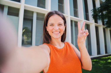 Güzel bir genç bayanın portresi dişlek bir gülümsemeyle üniversite kampüsünde selfie çekerken eğleniyor. Heyecanlı beyaz, büyüleyici, mutlu kadın dışarıda barış işareti gösteren kameraya bakıyor.