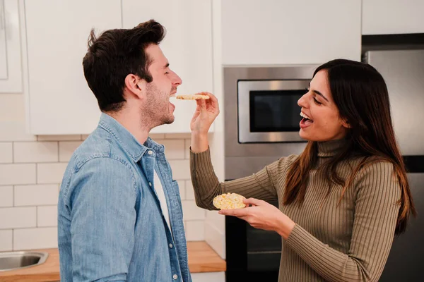 Young couple eating together in affeccionate attitude. Two people laugh and feed each other at home kitchen. Smiling wife flirting giving healthy food to her handsome husband having a romantic lunch