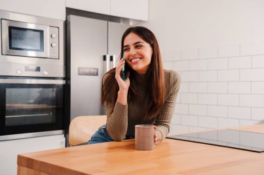 Genç bir kadın cep telefonuyla arıyor. Ev mutfağında gülümseyen ve akıllı telefondan konuşan mutlu genç bir bayan. Esmer, neşeli bir kadın dairesinde cep telefonuyla dostça sohbet ediyor.