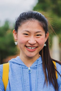 Kampüste dikilen ve gülen Asyalı sevimli kızın dikey yakın portresi. Genç Çinli kız öğrenci lisede kameraya olumlu bir ifadeyle gülümsüyor. Yüksek kalite