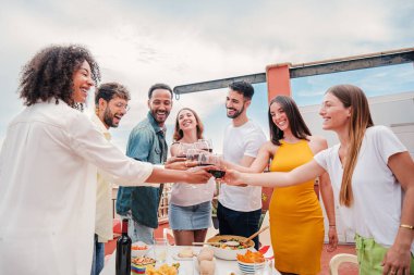 Group of multiracial adult friends smiling and clinking glasses of wine on a rooftop dinner party. Young happy people having fun toasting with alcohol celebrating together on a friendly meeting. High clipart