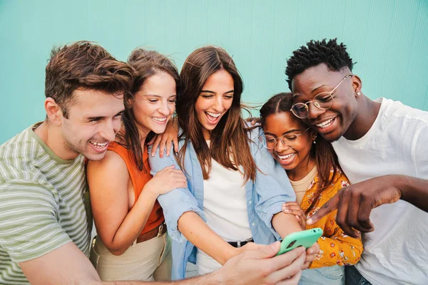 青い壁を背景に スマホアプリを使って楽しそうに笑顔を浮かべる若者たちの多人種グループ ソーシャルメディアでコンテンツを見ることを楽しんでいる多様な10代の学生 — ストック写真
