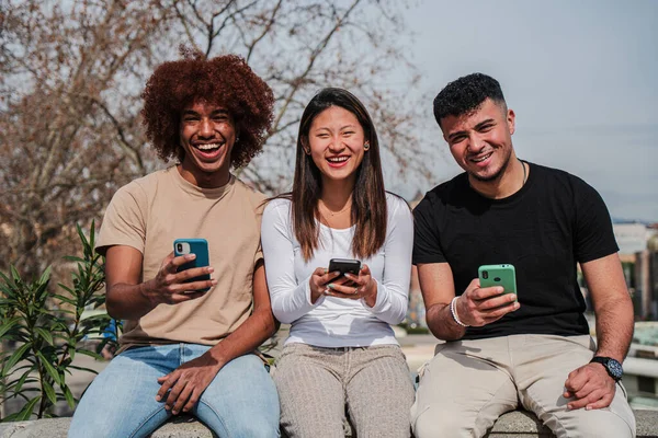 屋外で携帯電話デバイスを使用して楽しんでいる多人種の十代の友人のグループ 3人の多民族の若者が路上でソーシャルメディアで携帯電話をシェアするのを見て笑って笑っています — ストック写真