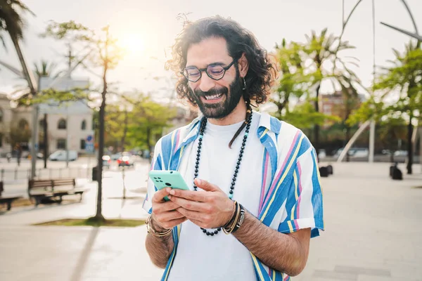 Genç yetişkin bir adam sokakta yürürken akıllı telefon mesajlarını ve sohbetlerini kullanarak boş zamanlarının tadını çıkarıyor. Cep telefonu tutan, mesaj atan ya da sosyal medya uygulamasında gezinen gülümseyen adam.