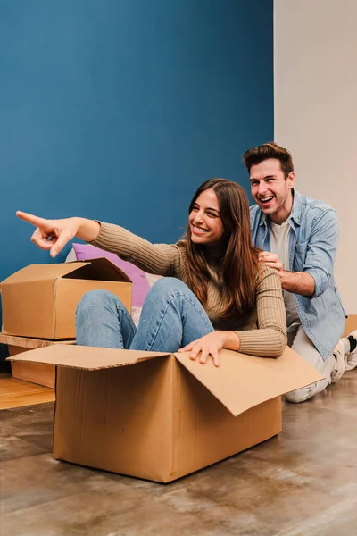 Yeni dairelerinde bavullarını açarken oynayan bir çiftin dikey portresi. Karton bir kutuda oturan mutlu kadın, yeni evine taşınırken gülümseyen erkek arkadaşıyla eğleniyor.