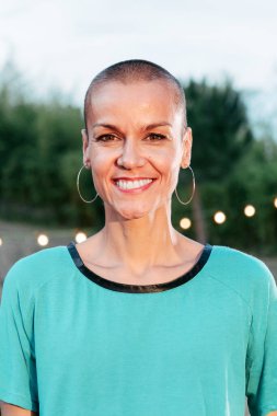 Kısa saçlı, gülümseyen ve kameraya bakan modern bir kadının dikey bireysel portresi. Yetişkin, mutlu bir dişi önümüze bakıyor. Havalı neşeli bayan gecenin karanlığında evin arka bahçesinde dikiliyor.