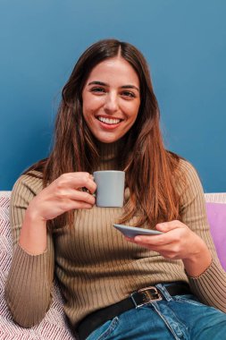 Kafkasyalı genç mutlu bir kadının dikey portresi elinde kahve fincanı, gülümsüyor ve rahat dairesinde kanepede oturmanın tadını çıkarıyor. Neşeli kadın evde çay içiyor.