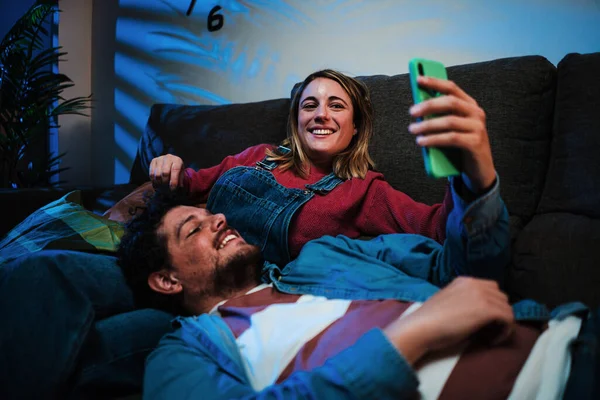 Cheerful couple using smart phone resting on couch at night. Smiling boyfriend leaning on his girlfriend legs watching at his cellphone watching social media relaxing and enjoying the weekend at home