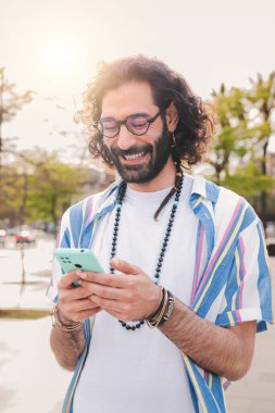 Akıllı telefon mesajlarını kullanan, sohbet eden, yürüyen ve boş zamanlarının tadını çıkaran genç bir adamın dikey portresi. Cep telefonu tutan, mesaj atan ya da sosyal medya uygulamasında gezinen gülümseyen bir adam. Yüksek