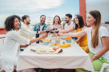 Sağlıklı bir yemek ve kırmızı şarap kadehleri eşliğinde kutlama yapan mutlu, çok kültürlü yetişkin arkadaşlar grubu. Arkadaşlar öğle yemeği şöleninde eğleniyor ve tezahürat yapıyorlar.