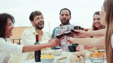 Sağlıklı bir yemek ve kırmızı şarap kadehleri eşliğinde kutlama yapan mutlu, çok kültürlü yetişkin arkadaşlar grubu. Arkadaşlar öğle yemeği şöleninde eğleniyor ve tezahürat yapıyorlar.
