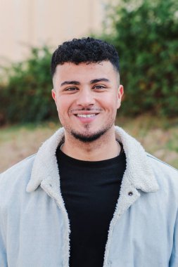 Gülümseyen ve kameraya bakan genç adamın dikey portresi. Üniversite kampüsünde dışarıda duran genç bir erkeğin ön görüntüsü. Öğrenci çocuk liseye gülüyor. Genç adam suratı. Yüksek