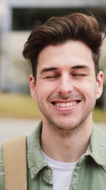 Üniversite kampüsünde beyaz, mutlu bir erkeğin portresini dikey olarak yakın çekiyoruz. Üniversitede ya da lisede durup gülen, gülümseyen ve kameraya bakan genç bir öğrencinin portresi. Yüksek