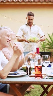 Arka bahçedeki öğle yemeği partisinde ailesiyle sağlıklı yiyecekler paylaşan yakışıklı, orta yaşlı bir adamın dikey portresi. Yüksek kaliteli görüntüler