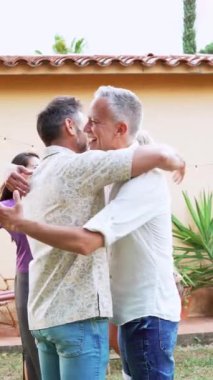 Dışarıdaki sosyal bir toplantıda, arka bahçedeki olgun bir arkadaş grubu arasındaki dostça selamlaşmanın dikey portresi. Orta yaşlı insanlar birbirlerine sarılıyorlar. Yüksek kaliteli görüntüler