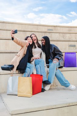 Alışveriş yaptıktan sonra cep telefonuyla selfie portresi yaparken gülümseyen bir grup çok ırklı genç kadının dikey portresi. Üç çoklu mutlu kız dışarıda oturup fotoğraf çekerken eğleniyor. Yüksek