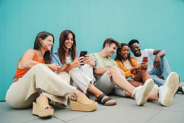 Çok ırklı gülümseyen gençler sosyal medyada telefonlarıyla mavi arka planda otururken eğleniyorlar. Genç çoklu etnik öğrenciler eğleniyor ve gülüyorlar.