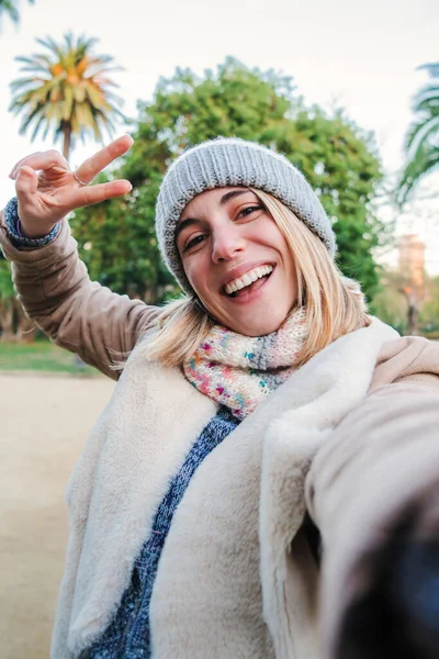 Vertical individual portrait of one happy caucasian blonde girl smiling taking a selfie portrait with a cellphone. European happy young woman doing the peace sign with the fingers standing outdoors