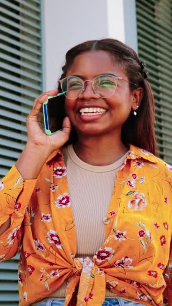 一个非洲裔美国女学生用智能手机说话的垂直肖像 一位惊慌失措的女人一边站在室外打电话 一边在友好的交谈中笑着 — 图库视频影像