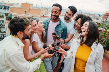 Bir grup neşeli genç arkadaş çatıdaki doğum günü partisinde gülümseyip kırmızı şarap kadehlerine kadeh kaldırıyorlar. Mutlu yetişkinler hafta sonu kutlamalarında kadeh tokuştururken eğleniyorlar. Arkadaşların elinde içki var.