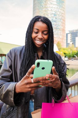 Genç bir Afrikalı Amerikalı kadının dikey portresi gülümseyen ve cep telefonu uygulaması ile daktilo eden. Açık havadan alışveriş yaptıktan sonra akıllı bir telefonla mesaj gönderen mutlu genç kız. İletişim