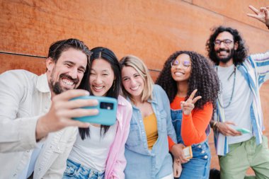 Bir grup yetişkin arkadaş selfie çektirip gülümsüyor ve birlikte gülüyorlar. Arkadaş canlısı insanlar dışarıda cep telefonuyla sosyal medya uygulamasında fotoğraf çekerken eğleniyorlar.