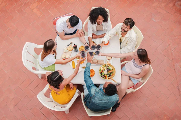High angle view of a group of friends having a dinner party celebration at home terrace, toasting or clinking red wine glasses. Social meeting of a young adult buddies sitting together at table. High
