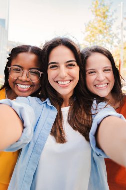 Bir grup kızın dikey portresi. Mutlu çok ırklı genç kızlar birlikte selfie çekerken gülümsüyorlar. Çok ırklı genç bayan arkadaşlar gülüyorlar ve akıllı telefonla fotoğraf çekiyorlar.