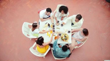 Bir grup arkadaşın evdeki terasta kutlama yaparken, kadeh kaldırırken ya da kırmızı şarap bardaklarını tokuştururken yüksek açılı görüntüsü. Masada birlikte oturan genç bir yetişkinin sosyal toplantısı. Yüksek