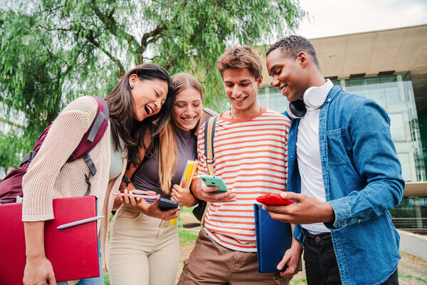 Group of students having fun together sharing on the social media with a cellphone app. Happy friends using their smartphones. Multiracial teenagers browsing on internet with a mobile phone at campus