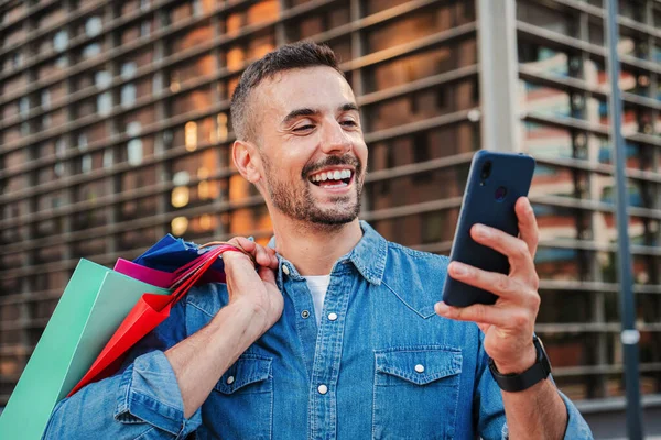 Portrait of handsome happy guy purchasing a sale online on a marketplace with a cellphone app holding shopping bags. Shopaholic man smiling searching a discount using a smart phone on a web store