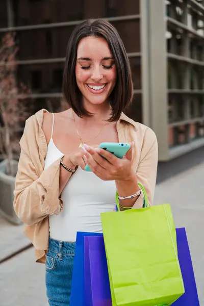 Vertical portrait of a young adult joyful woman using a cellphone to purchase online on a market place app. Smiling female carrying shopping bags and having fun watching a sale on a mobile phone. High