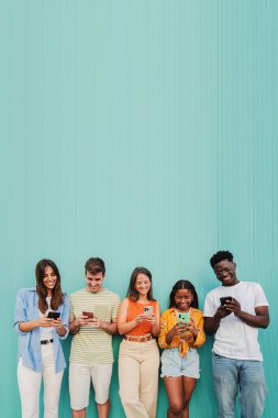 Dikey. Teal Wall 'daki cep telefonu uygulamalarını kullanarak gülümseyen çok ırklı bir grup genç arkadaş. Gençler sosyal medyada mavi arka planda cep telefonuyla mesajlarını paylaşırken eğleniyorlar. Yüksek
