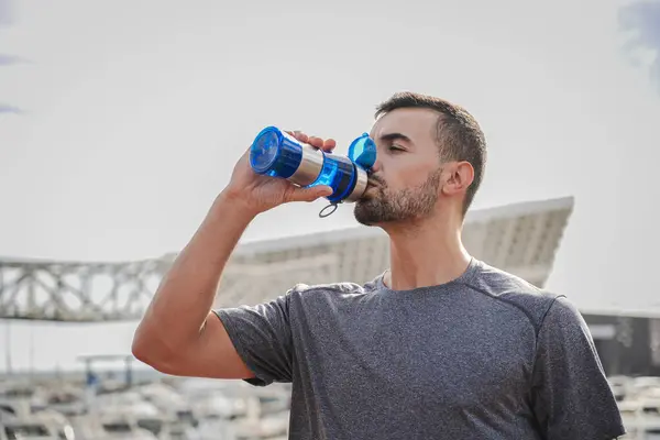 若いハンサムなスポーツマンは ランニングやトレーニング後に休んでいる間 ボトルからエネルギー水を飲んでいます 屋外トレーニング中にサプリメントとさわやかな休憩を持つ強い複合体の男性 — ストック写真