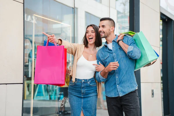 Happy couple enjoying a shopping day together, watching for sales, retails, promotions and discounts in the family mall. Pretty woman pointing a store showcase and her boyfriend carrying purchase bags