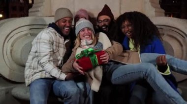 Heyecanlı genç bir kadın kollarında doğum günü hediyesi ya da en iyi arkadaşlarının birlikte güldüğü bir Noel hediyesi tutuyor. Mutlu bir grup çok ırklı insan Noel gecesini kutluyor.
