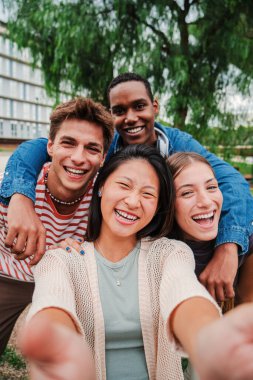 Bir grup genç çok ırklı lise öğrencisinin dikey portresi dişlek bir gülümsemeyle dışarıda kameraya bakıp gülüyor ve birlikte eğleniyorlar. Arkadaş canlısı gençler fotoğraf çekiyor.