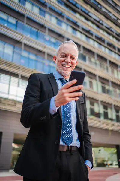 Vertical. Middle aged businessman using a cellphone to watch his finances outside. Mature business man sending messages with a smartphone app. White collar worker smiling and holding a mobile phone