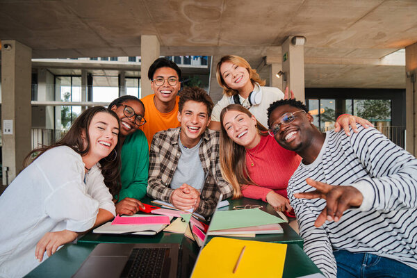 Happy young university students smiling and looking at camera enjoying together sitting in the library or classroom. Multiracial teenage friends laughing on a study break at campus. High school people