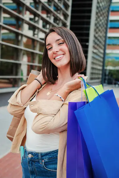 Vertical portrait of a young woman holding shopping bags and looking excited at camera. Joyful customer female enjoying a discount purchase. Lady smiling buying a retail. Buyer girl at sale store