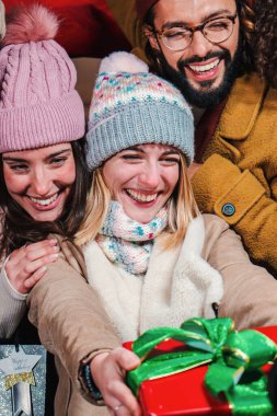 Heyecanlı bir kadının arkadaşına Noel Baba hediyesi verirken dikey portresi. Geleneksel Noel hediyesini veren neşeli bir kadın. Doğum gününü kutlamak için paketlenmiş bir kutu tutan genç kız.