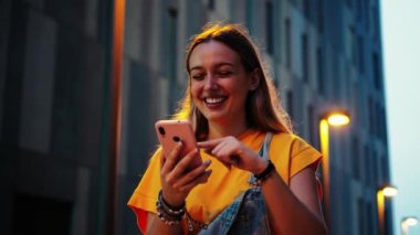 Geceleri cep telefonu kullanan genç bir kadın. Mutlu kadın, alacakaranlıkta akıllı telefon sosyal medya uygulaması ile eğleniyor. Genç bir kız internette cep telefonu uygulaması ile geziniyor.