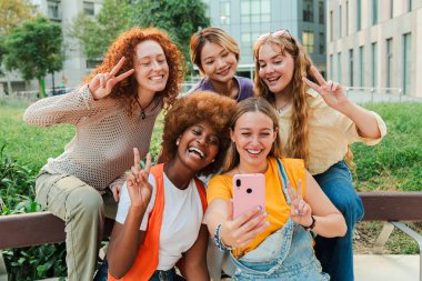 Modern bir akıllı telefonla kameraya bakan mutlu, çok ırklı genç kadınlar dışarıda bankta otururken eğleniyorlar. Komik kadınlar, arkadaşlarının cep telefonlarıyla selfie çekerken eğleniyorlar.