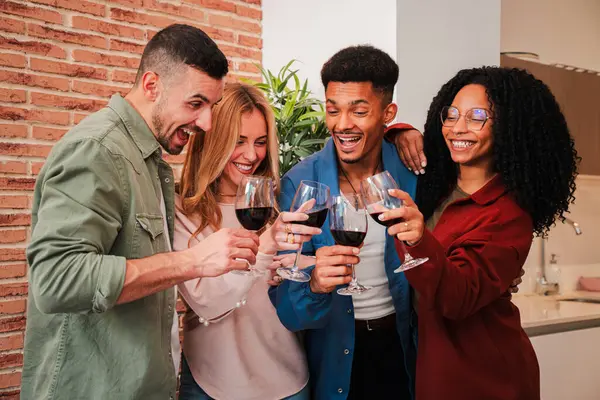 Group of young adult friends having fun toasting a red wine glasses at home reunion or birthday party. People drinking alcohol. Multiracial couples enjoying on a social gathering celebrating together