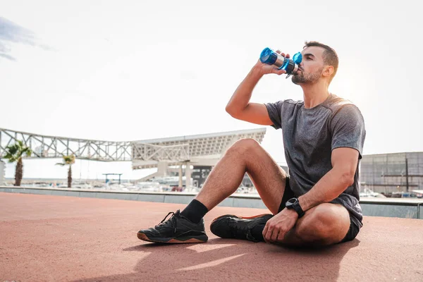疲れたスポーツマンは 床に座っている耐久ワークアウトの後に休んで水を飲む 疲労した男性は回復し 心臓ウェルネストレーニングでタンパク質サプリメントを取る定期的な休憩を持っています — ストック写真