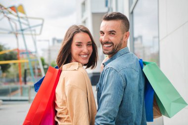 Alışveriş merkezinde alışveriş torbaları taşıyan genç bir çiftin arka görüntüsü. Neşeli erkek arkadaş ve mutlu kız arkadaş satış gününde mağazadaki kameraya bakıyor. Mağazadan alışveriş yapan heyecanlı insanlar