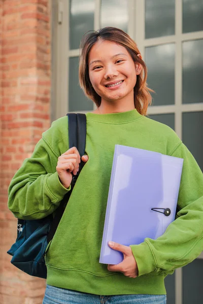 Vertical portrait of a young asian high school student girl smiling and looking at camera. Chinese teenage female staring front at university campus. Thai classmate woman with happy expression. High