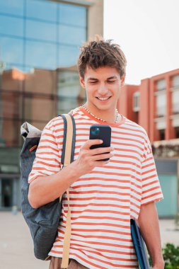 Dikey. Erkek öğrenci kampüsteki sosyal medyada cep telefonuyla eğleniyor. Gerçek bir genç adam akıllı telefon kullanarak gülümsüyor. Elinde cep telefonu olan genç bir adam. Genç okul çocuğu telefon okuyor.