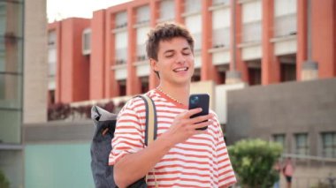 Erkek öğrenci üniversite kampüsündeki sosyal medyada cep telefonuyla eğleniyor. Gerçek bir genç adam akıllı telefon kullanarak gülümsüyor. Elinde cep telefonu olan genç bir adam. Genç okul çocuğu telefon okuyor.